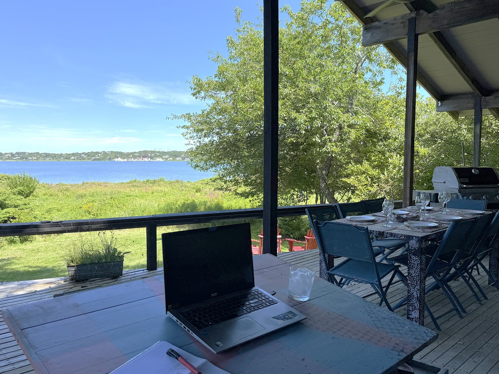 outdoor covered deck showing Weber propane gas grill, dining table, and a drawing board used as a workspace, with Lake Montauk in the background